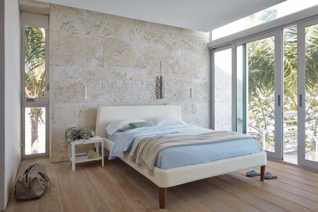 A modern bedroom featuring a minimalist design with a white upholstered bed, light blue bedding, a side table with a plant, and large glass doors revealing a view of palm trees outside.