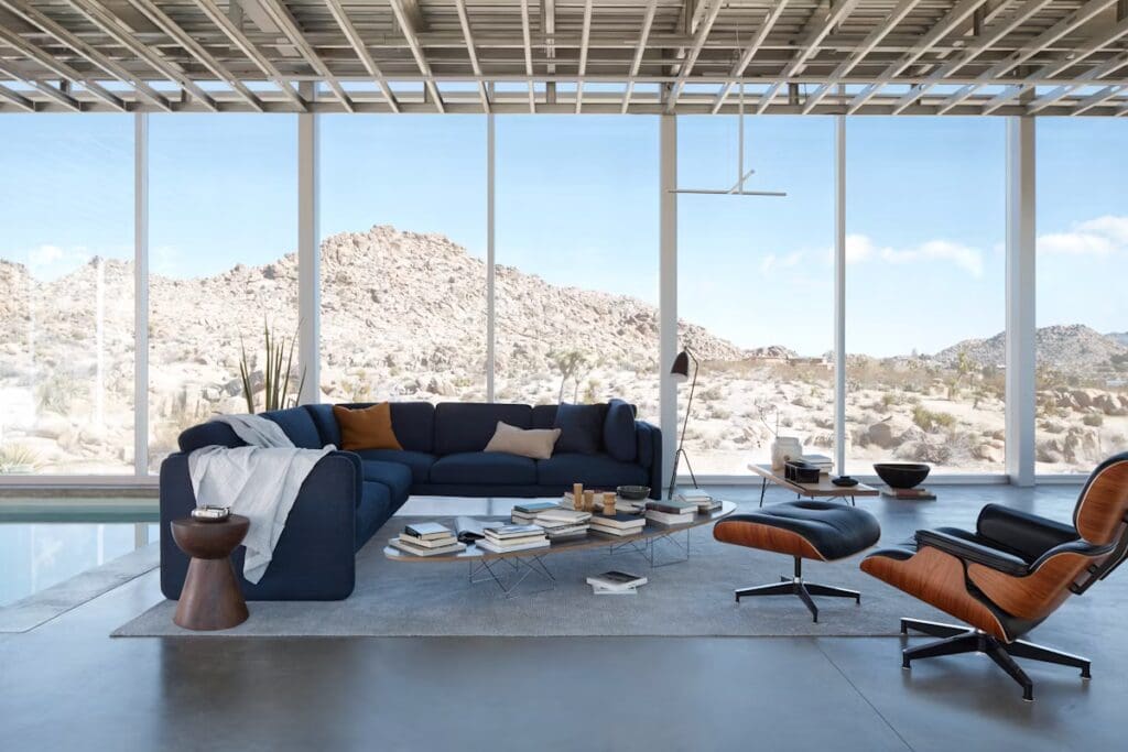 A modern living room featuring a navy blue sectional sofa, a low coffee table with stacked books, a stylish accent chair, and large glass windows providing a view of a rocky landscape.