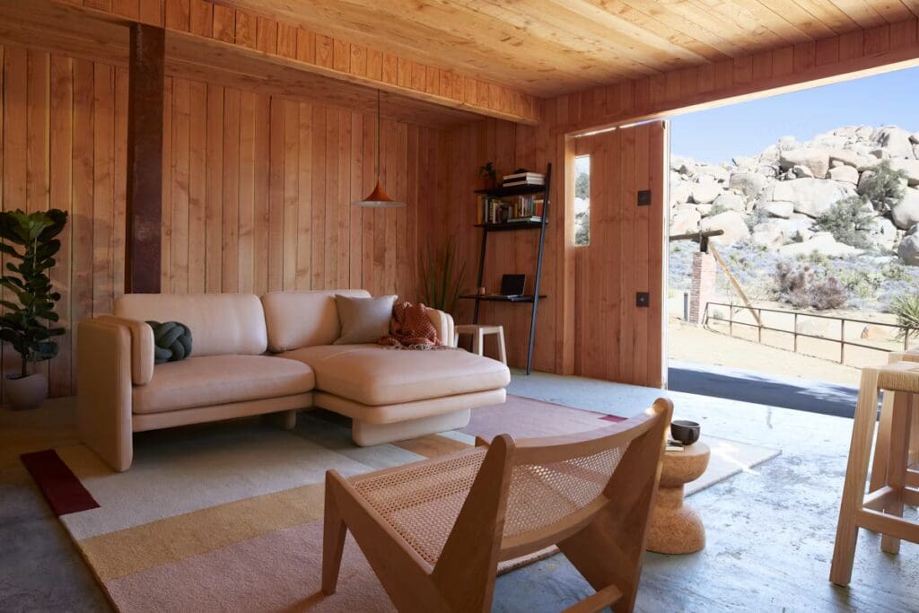 A modern living room with a light-colored sectional sofa, a patterned area rug, and wooden walls and ceiling. A potted plant adds greenery, while a wooden chair and side table complement the design. Large windows offer a view of a rocky landscape outside.