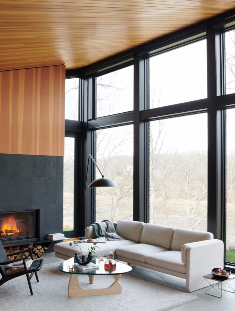 A modern living room with a light-colored sectional sofa, a round glass coffee table, a black lounge chair, and a stylish pendant lamp, featuring large windows with a view of nature outside and a cozy fireplace.