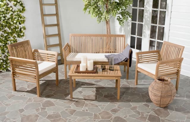 Modern outdoor seating set with wooden chairs and a bench, featuring plush white cushions, a coffee table with candles, and a decorative woven basket. A leafy tree and a white wall with windows are in the background.