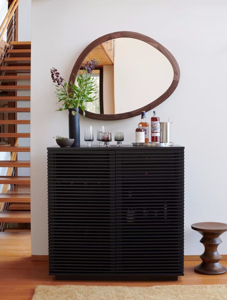 A modern living room featuring a stylish dark wooden cabinet with rounded mirror, a decorative vase with flowers, and liquor bottles set on the cabinet.