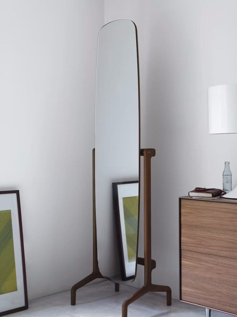 A tall, free-standing mirror with a wooden frame, accompanied by framed artworks leaning against the wall and a wooden dresser nearby.