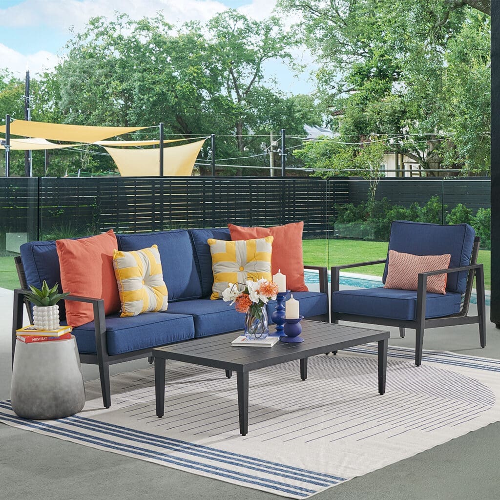 Stylish outdoor living space featuring a navy blue sofa with colorful cushions, a matching armchair, and a decorative coffee table. The setting includes potted plants and a backdrop of greenery, creating a vibrant atmosphere.