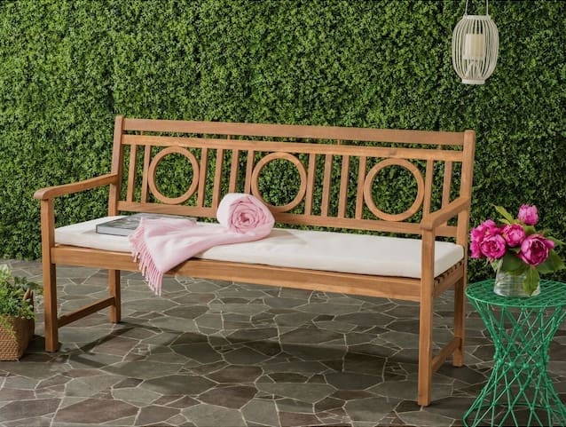 Modern wooden bench with a decorative backrest, featuring a pink throw blanket and a book on the seat, placed against a lush green wall and beside a vibrant flower arrangement.