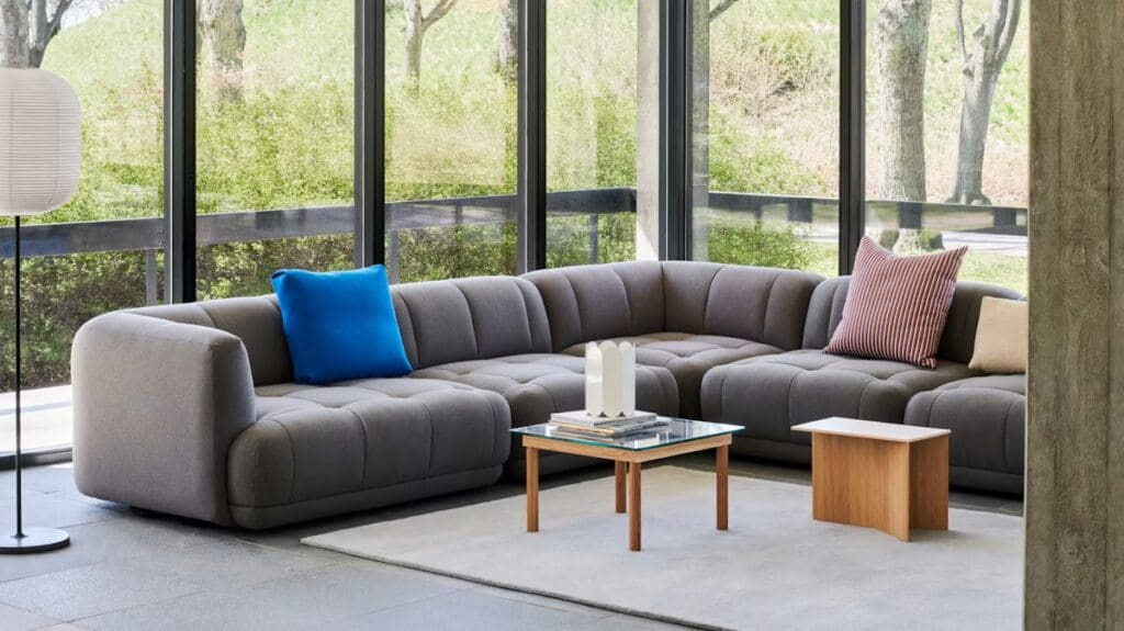 A modern living room showcasing a gray sectional sofa with a blue and red accent pillow, paired with a wooden coffee table and floor lamp, featuring large glass windows letting in natural light and a view of greenery.