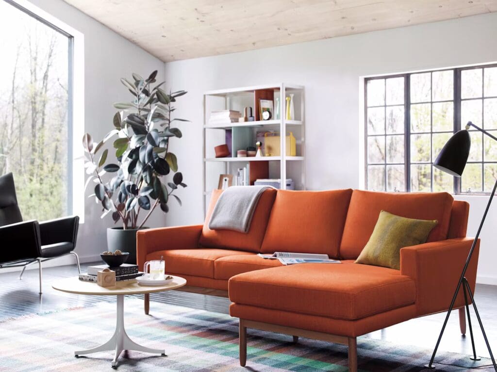 A modern living room featuring an orange sectional sofa with a cozy throw, a round wooden coffee table, and a stylish floor lamp. Large windows allow natural light to flood the space, complemented by a decorative plant and a colorful rug.
