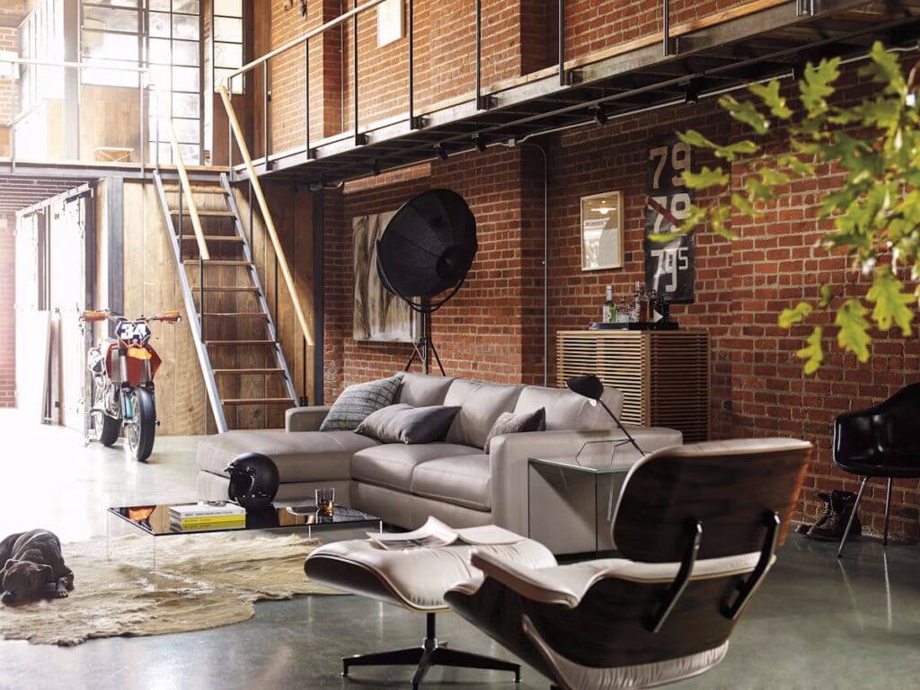 A modern living room with a gray sectional sofa, coffee table, stylish chairs, and industrial decor elements, featuring brick walls and a staircase.