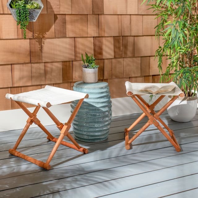 Two folding outdoor stools with a light cushion, placed near a decorative green vase with plants, against a wooden wall and surrounded by potted greenery.
