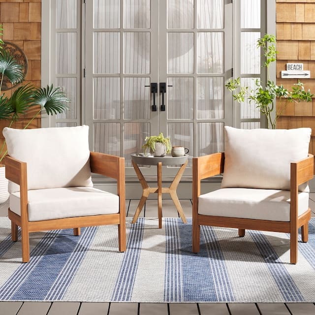 Cozy outdoor seating area featuring two light-colored armchairs with wooden frames, accompanied by a small round table. The setting is styled with plants and a patterned rug, creating a cheerful ambiance.