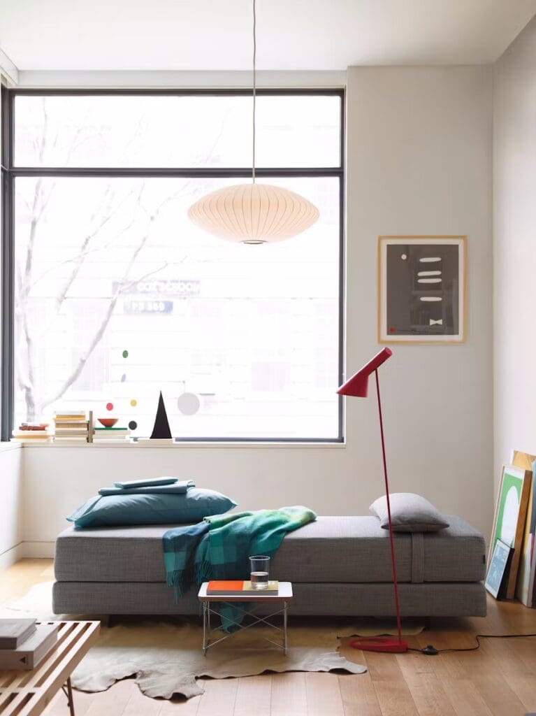 A modern living room scene featuring a gray sofa with cushions and a blanket, a red floor lamp, and a coffee table with a glass and a decorative item. There is a large window with natural light and contemporary art on the walls.
