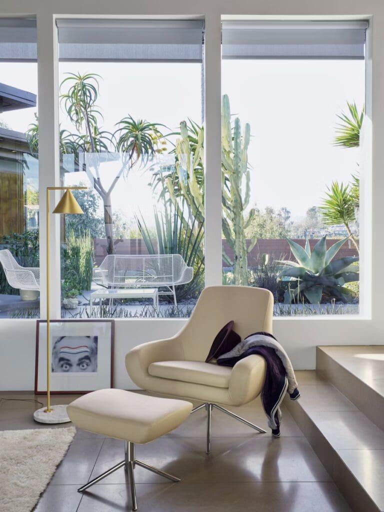 A modern living room featuring a sleek beige lounge chair with an ottoman, accompanied by a floor lamp. The large windows showcase a lush outdoor garden filled with various plant species.