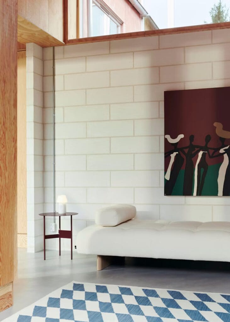 A modern interior featuring a white sofa, a round side table with a lamp, and a large piece of abstract art on the wall, set against white brick walls and wooden accents.