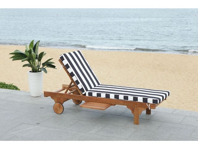 Wooden lounge chair with black and white striped cushions, positioned near a sandy beach with a view of the ocean and a potted plant beside it.