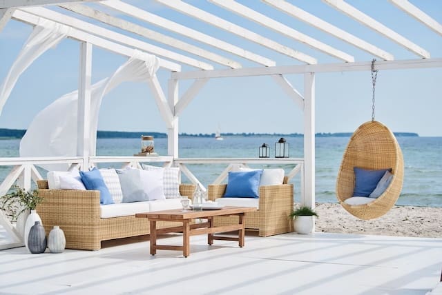 Stylish outdoor lounge area featuring a wicker couch with blue and white pillows, a wooden coffee table, and a hanging chair, all set against a beautiful beach and ocean view under a bright blue sky.