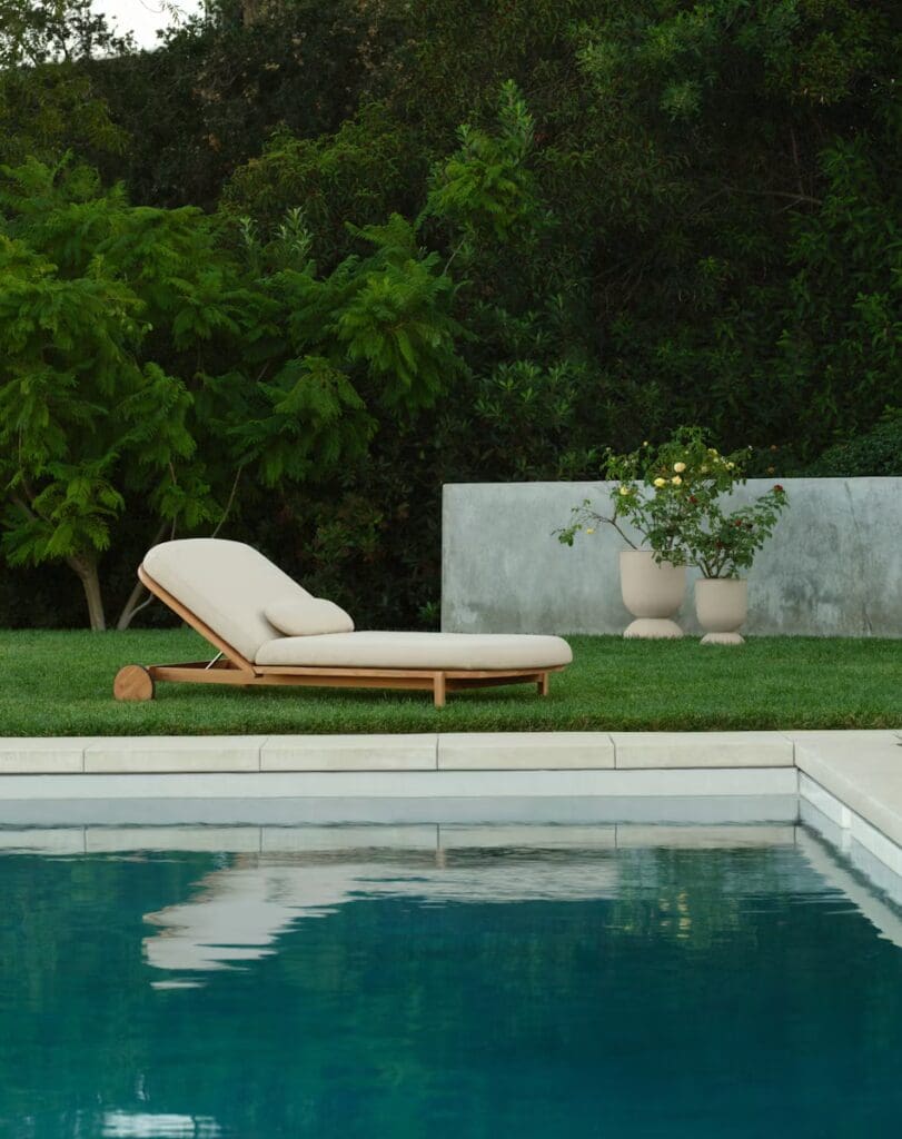 A modern chaise lounge positioned beside a pool, surrounded by lush greenery and a concrete wall, with a flowering plant in a decorative pot.