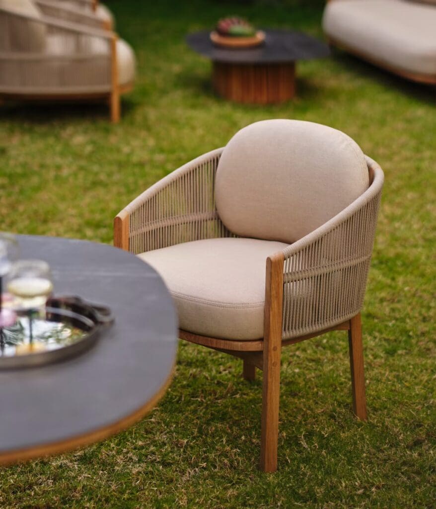 A stylish outdoor armchair with a rounded, cushioned seat and wooden armrests, set on a grassy area with a round table nearby.
