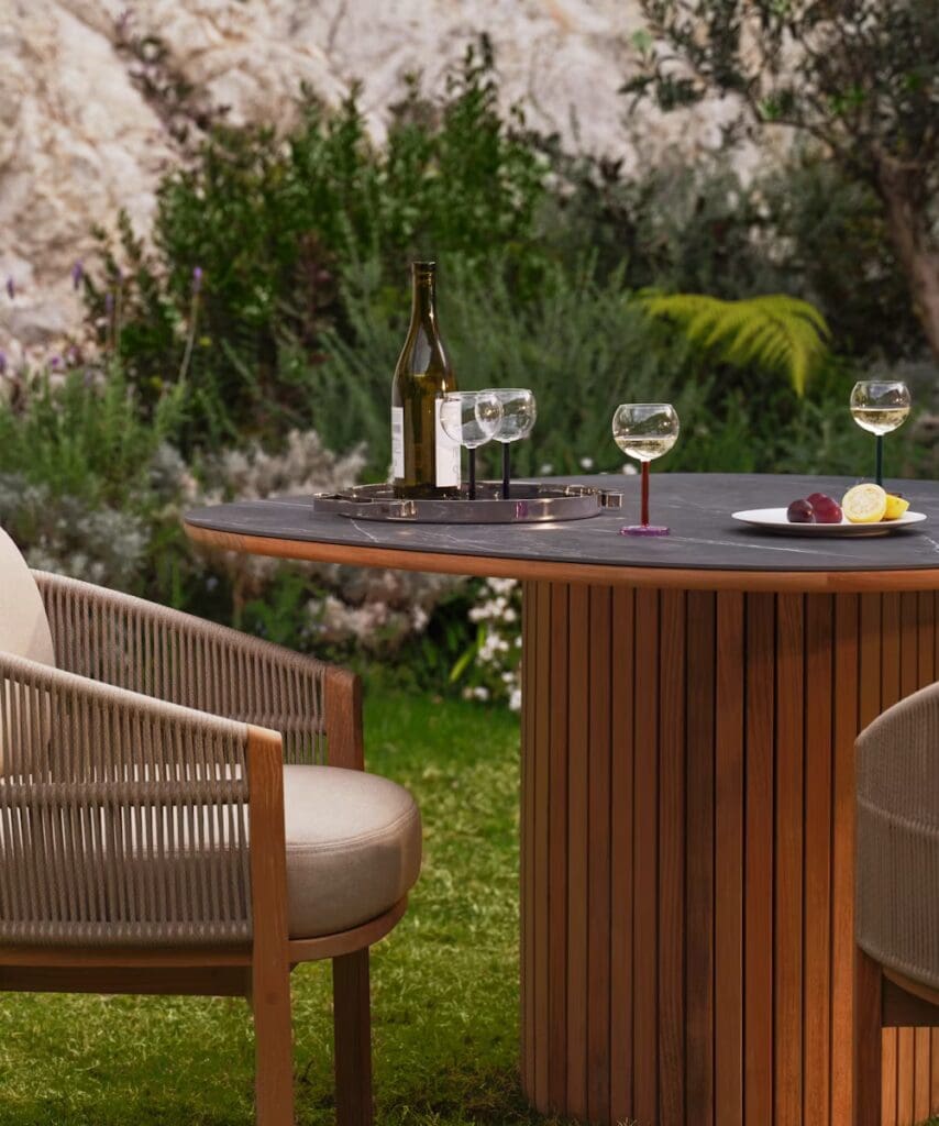 A close-up view of an outdoor dining setting featuring a round table with a dark surface, flanked by stylish chairs. On the table are a bottle of wine, three glasses, a small plate with lemon and grapes, and a serving tray, surrounded by lush greenery.