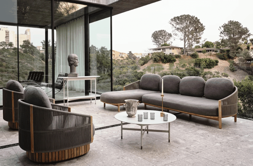 Stylish outdoor living space featuring a modern sectional sofa and armchairs with gray cushions, a round white coffee table, and a decorative bust, all framed by large glass windows and overlooking a hillside.