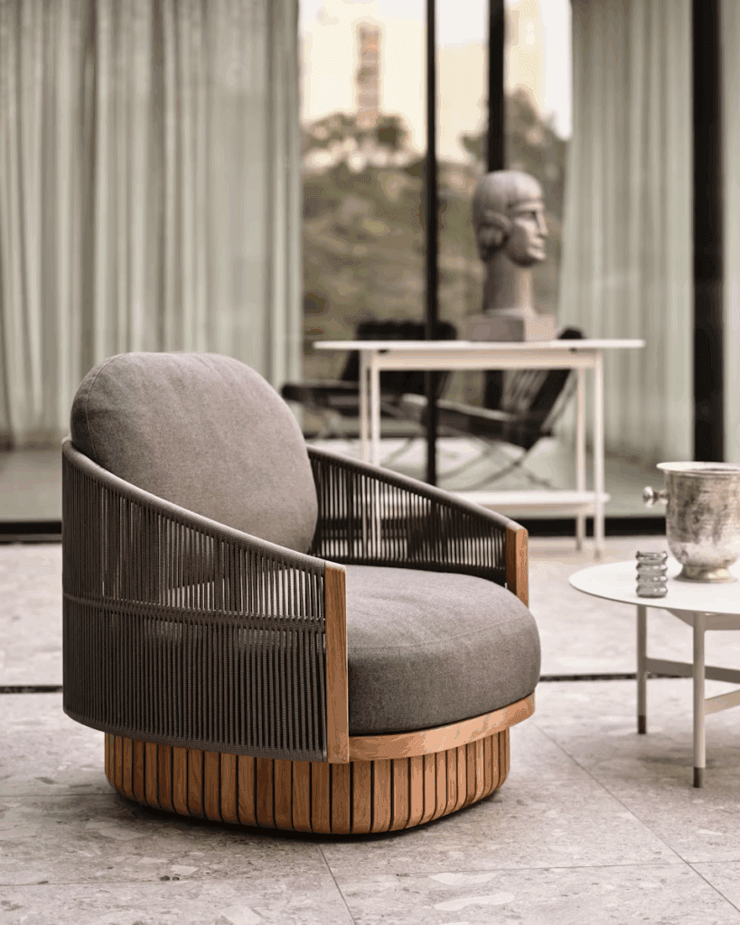 Modern outdoor chair with a rounded wooden base and fabric cushion, placed in an elegant setting with large glass windows and a minimalist table.