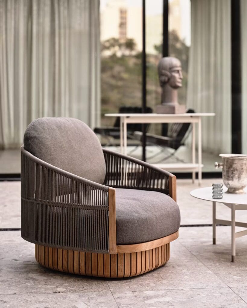 A modern lounge chair with a round base, featuring a soft grey cushion, wooden armrests, and a woven backrest, placed next to a small round table in an indoor space with large windows.