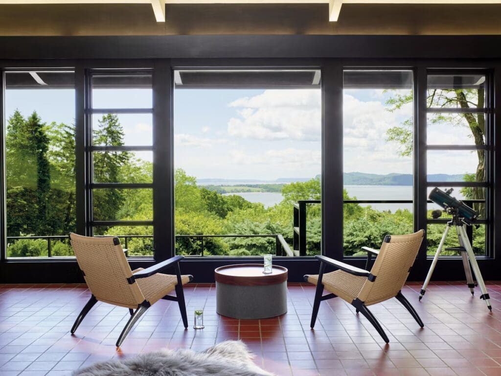 A modern living room with two woven chairs facing a round coffee table, large windows showcasing a lush outdoor view, and a telescope in the corner.