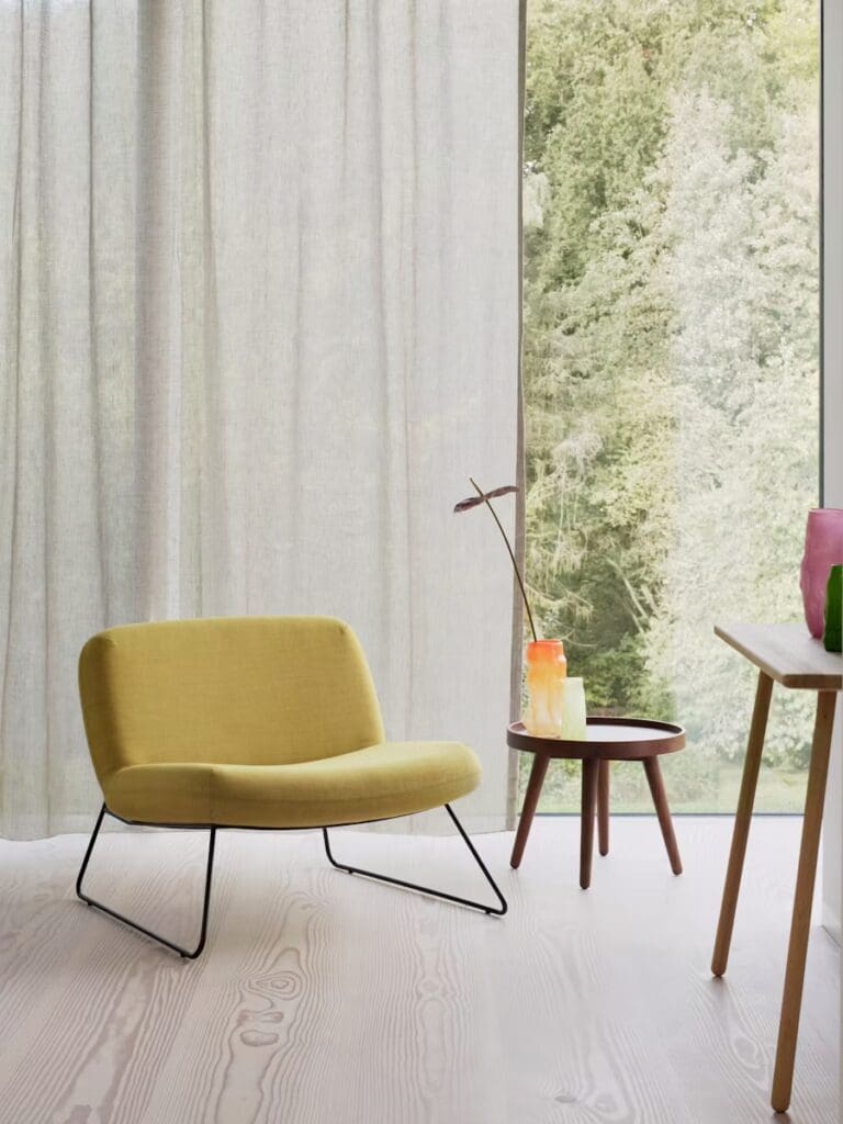 A modern living room scene showcasing a sleek yellow chair with a curved back and slim black legs. Next to it is a small round table with a vibrant glass sculpture, and in the background, sheer curtains filter natural light, revealing greenery outside.