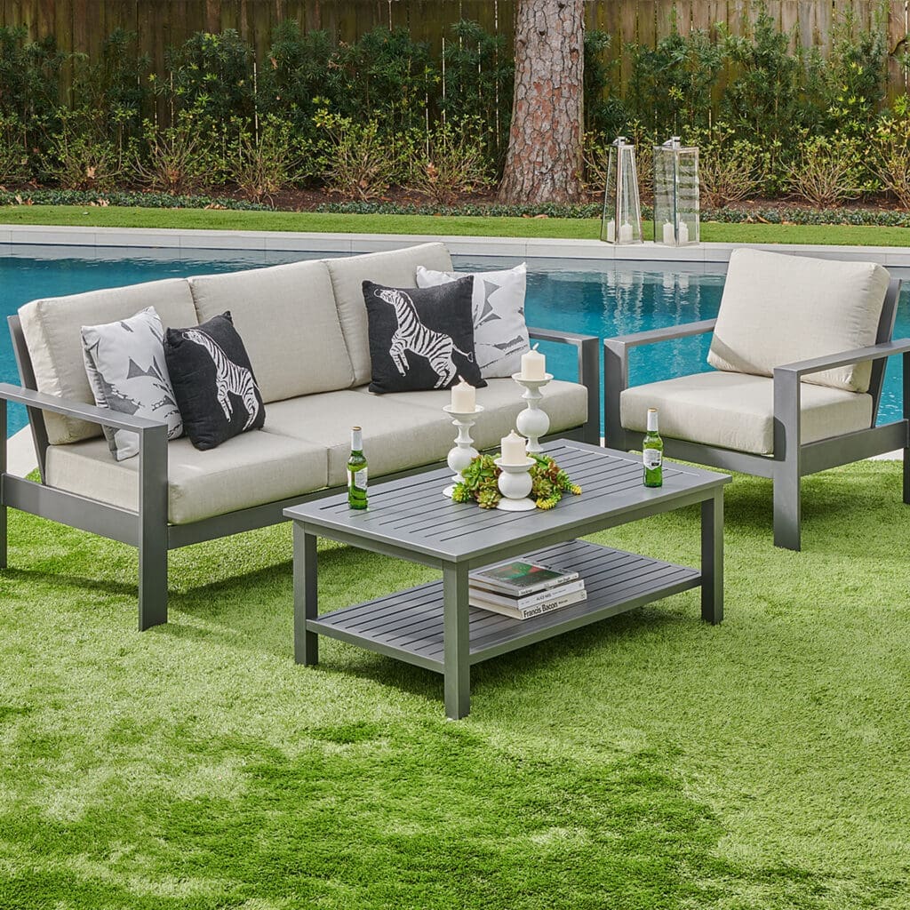 A stylish outdoor living space featuring a gray sofa with zebra-patterned cushions, a matching armchair, and a gray coffee table. The area is adorned with candles and a small succulent arrangement, set against a vibrant green artificial lawn and a reflective pool backdrop.