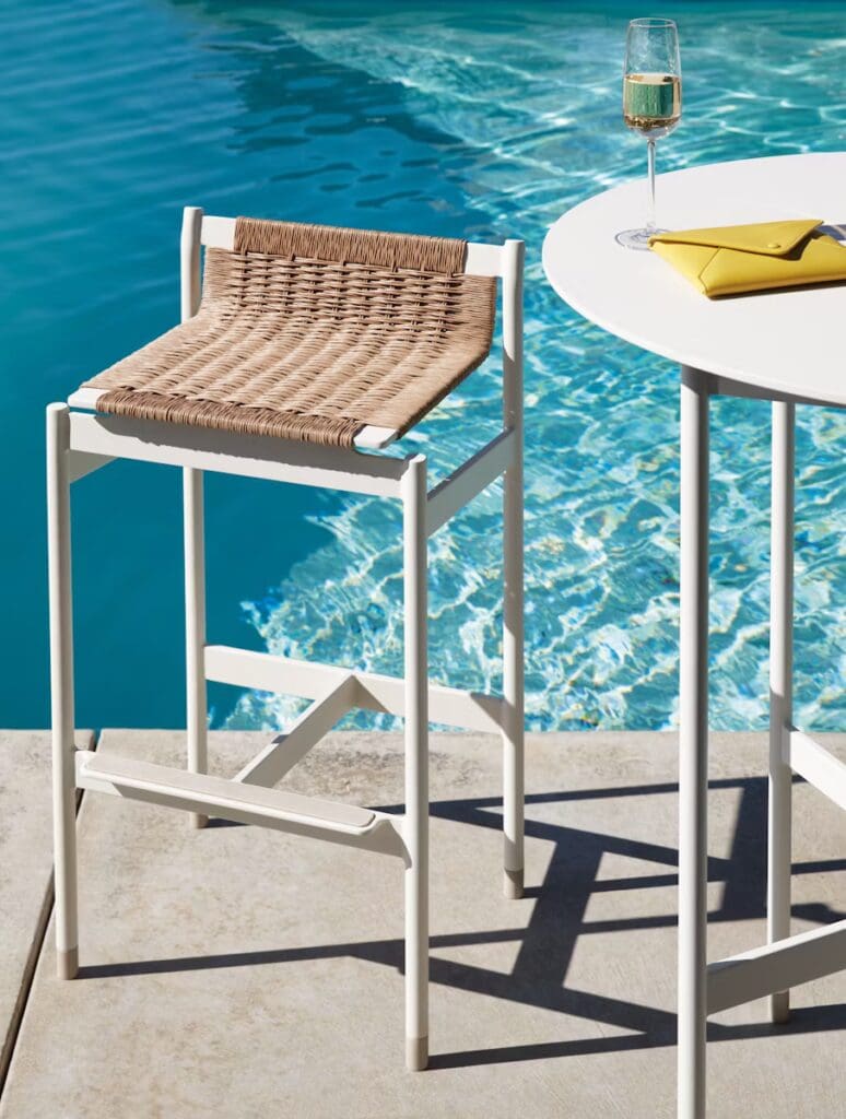 A stylish bar stool with a woven seat and a sleek white frame positioned next to a swimming pool, accompanied by a side table with a glass of champagne and a bright yellow clutch.