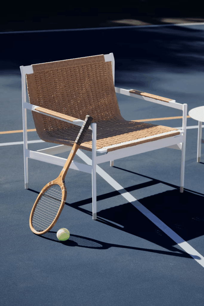 A modern wicker and white chair positioned next to a small table on a tennis court, with a tennis racket and ball resting on the ground.