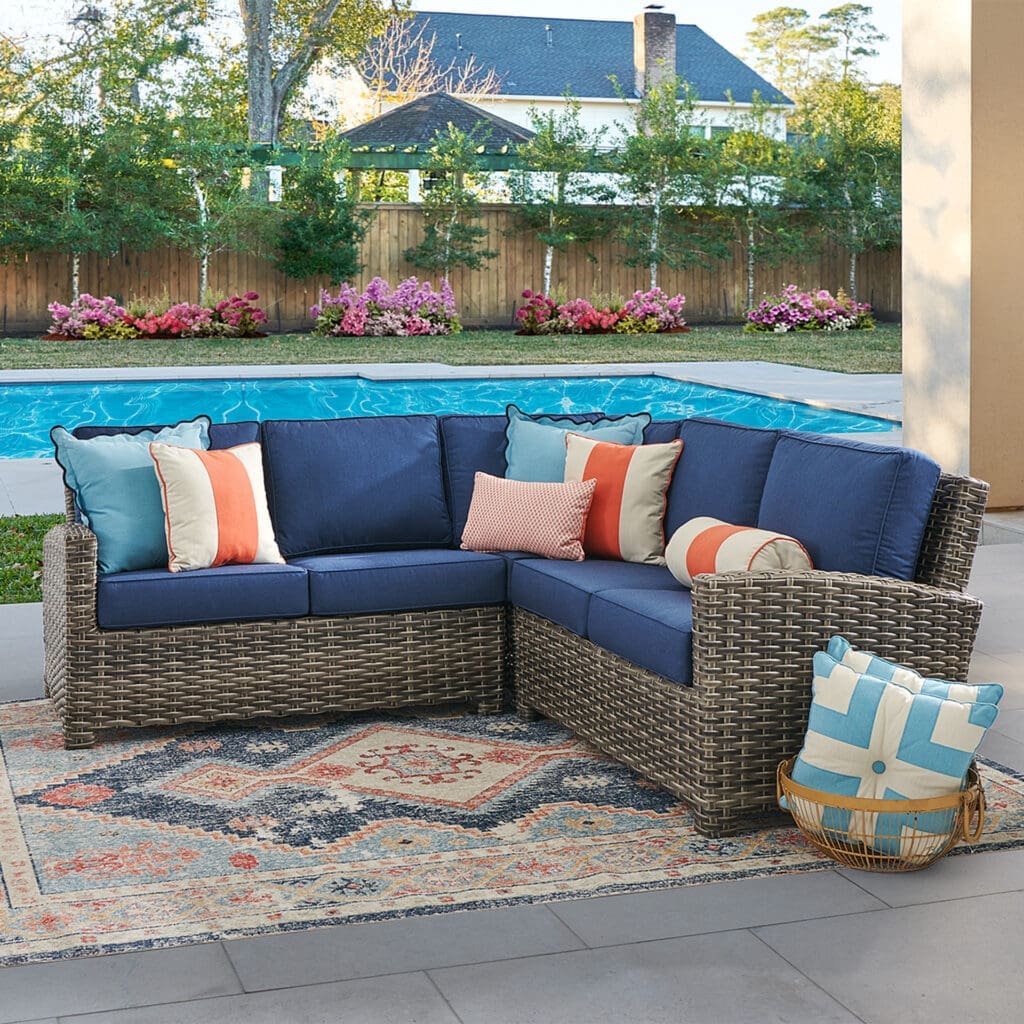 Cozy outdoor seating area featuring a sectional sofa with blue cushions and decorative pillows, placed on a patterned rug beside a sparkling pool.