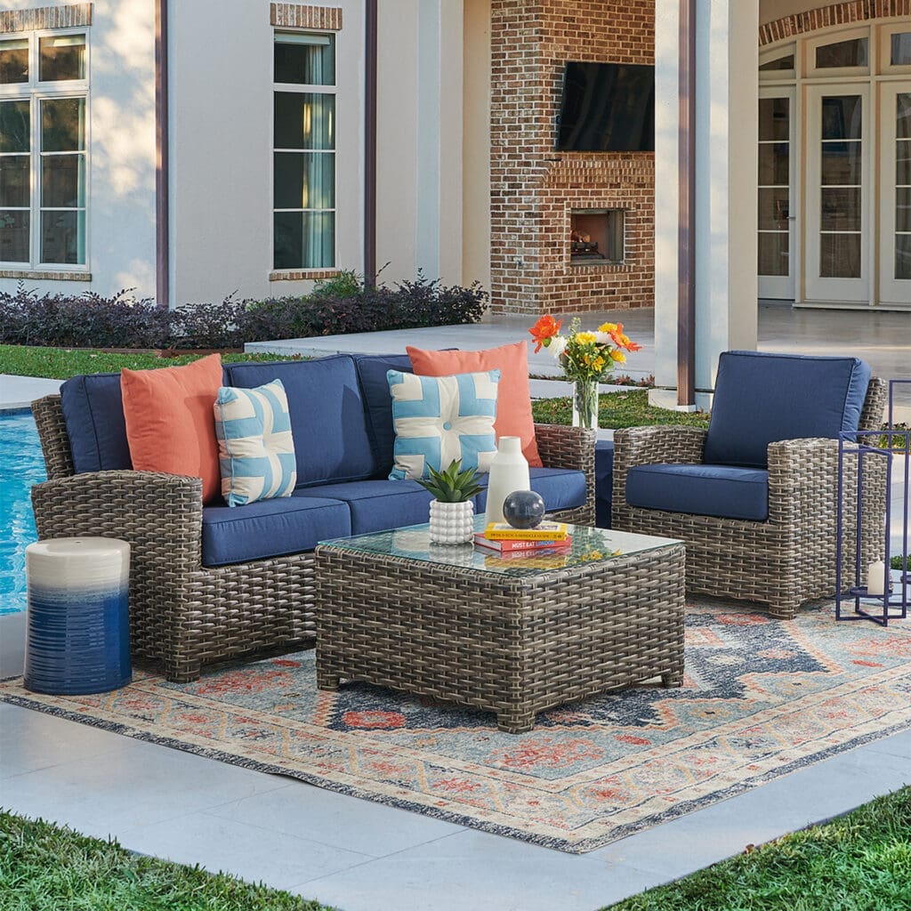 Cozy outdoor seating area with woven furniture, blue cushions, and decorative pillows beside a swimming pool, featuring a glass-top coffee table and a vibrant area rug.