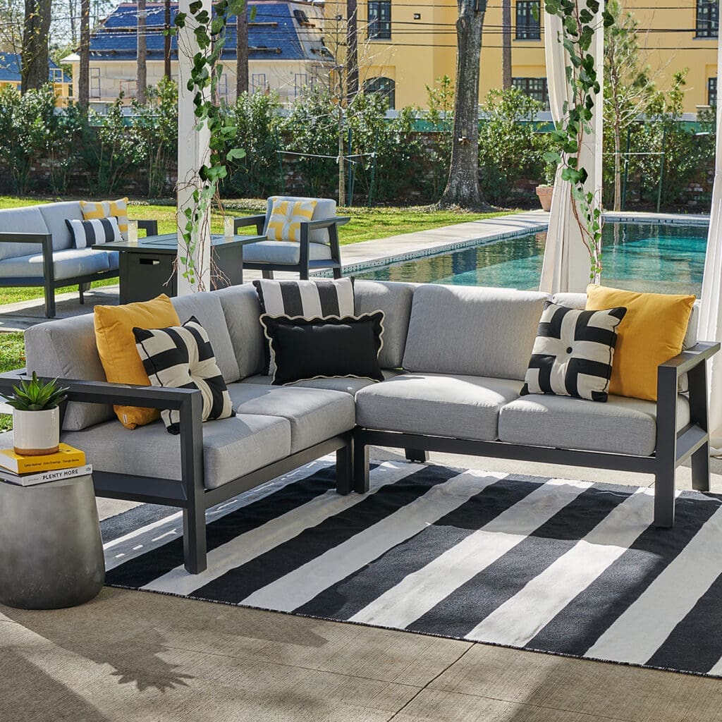 Luxurious outdoor seating area featuring a sectional sofa with grey cushions, decorative pillows, and a striped rug, overlooking a swimming pool and landscaped garden.