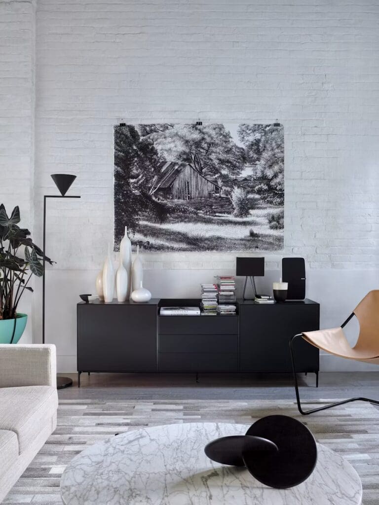 A modern living room featuring a light gray sofa, a marble coffee table with a round black decorative object, and a wooden sideboard with various decor items. A large black and white artwork depicting a rustic scene hangs on the white brick wall.
