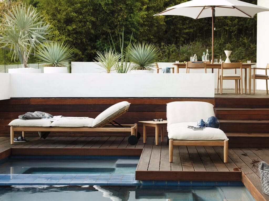 A stylish outdoor space featuring a wooden deck and a swimming pool, with a lounge chair, a cushioned seating area, and a dining table under an umbrella, surrounded by potted plants.