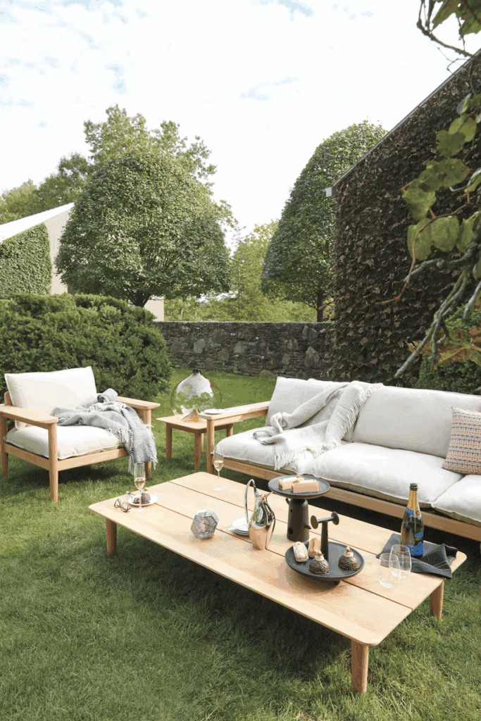 Stylish outdoor seating area with modern wooden furniture, featuring a comfortable sofa and armchairs, adorned with cushions and a cozy throw. A wooden coffee table displays decorative items and beverages, set in a lush green garden with trees.