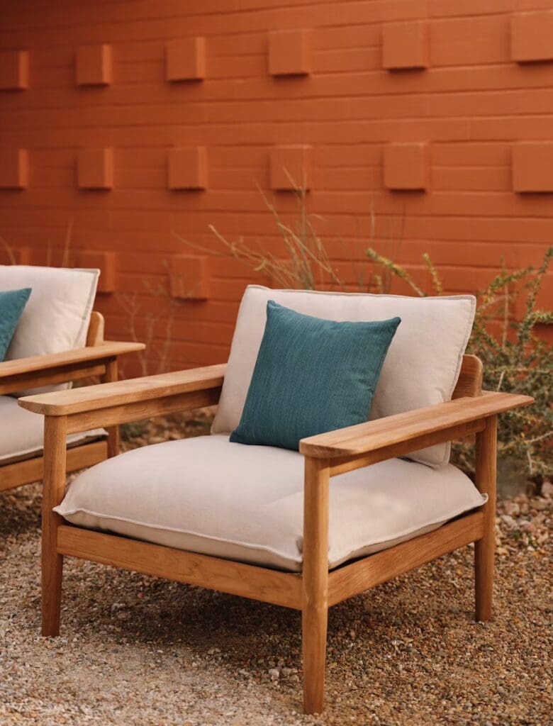 A stylish outdoor armchair made of wood with plush cushions and a teal accent pillow, set against a textured orange wall.