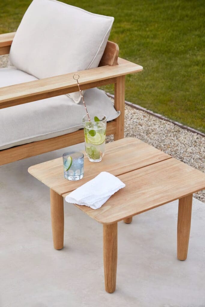 A modern outdoor setup featuring a wooden chair with a white cushion, accompanied by a wooden side table holding two glasses of drinks and a white napkin.