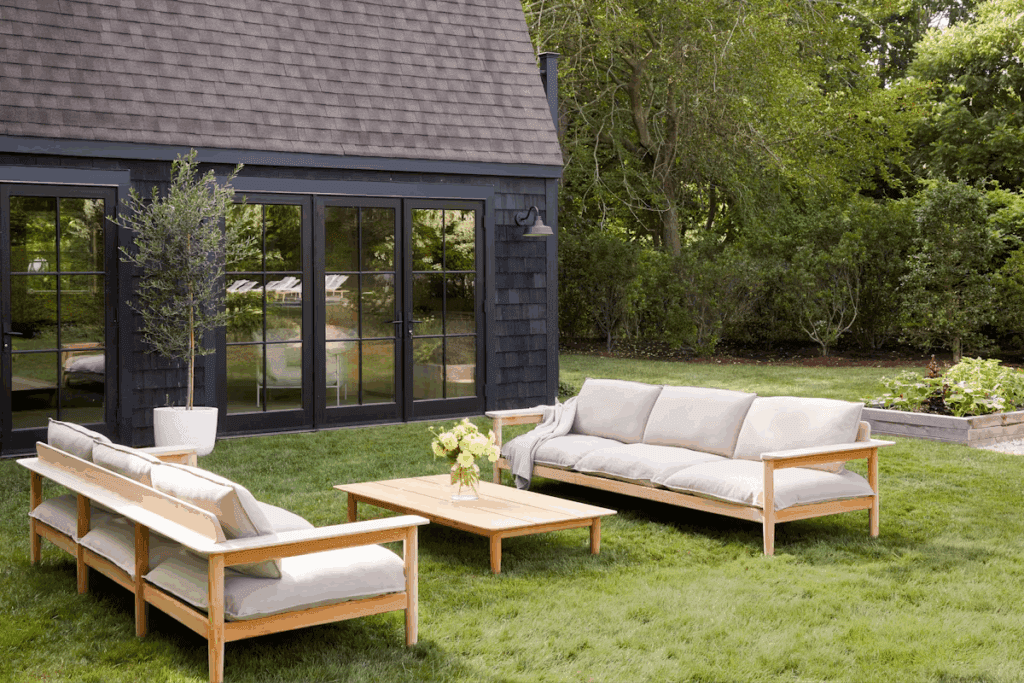 Outdoor living space featuring modern wooden furniture, including a sofa and coffee table, set on a lush green lawn next to a black house with large windows.