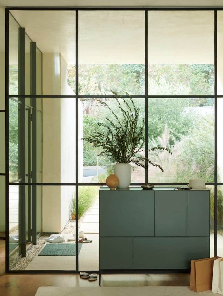 A modern entryway featuring a green storage cabinet, a vase with plants, and a large glass window revealing a view of the outdoors. The floor shows a welcome mat and shoes.