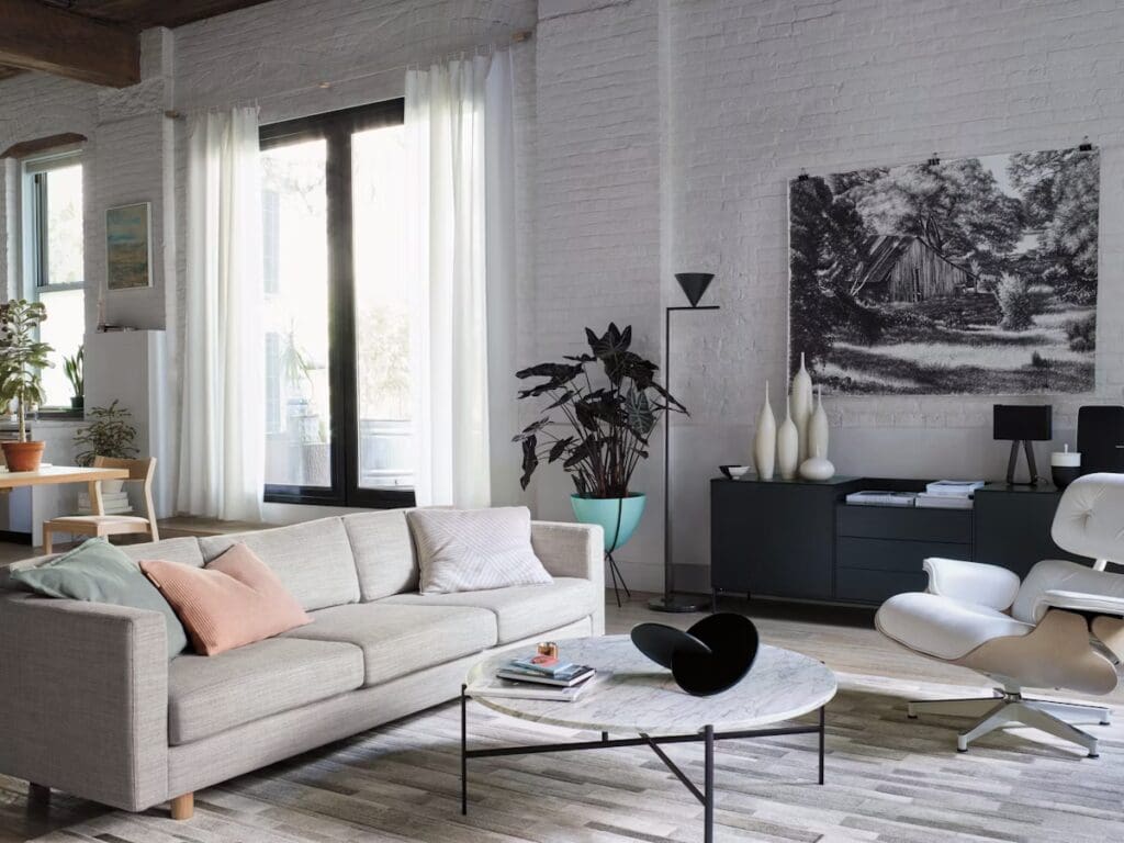 A stylish modern living room featuring a light-colored sofa, a round marble coffee table, and various decorative items. Large windows with sheer curtains allow natural light to fill the space, complemented by indoor plants and contemporary art on the walls.