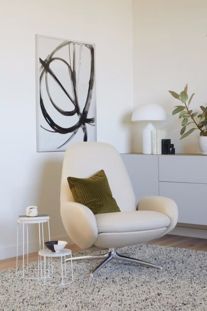A modern living room corner featuring a stylish cream-colored armchair with a green striped pillow, a small white side table with decorative items, a sleek lamp, and a potted plant. A contemporary abstract painting hangs on the wall beside light wood cabinetry.
