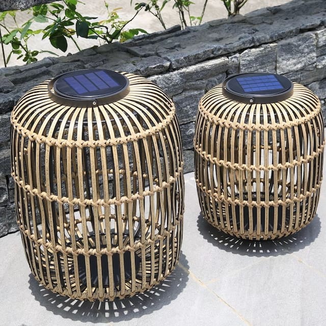 Two solar-powered outdoor lanterns with a woven bamboo design sitting on a stone surface, surrounded by greenery.