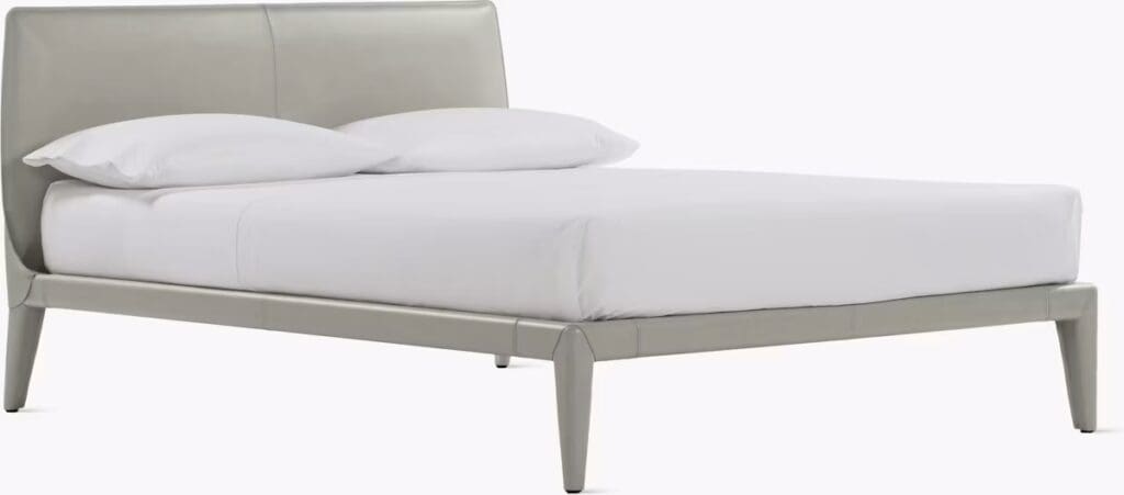 A modern bed featuring a sleek light gray leather frame with a contemporary design, complemented by white bedding and pillows.
