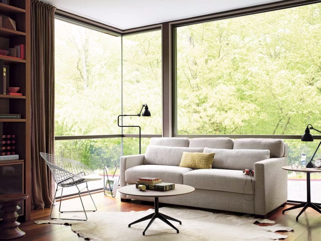 A modern living room with a light-colored sofa, a wooden coffee table, a decorative rug, and large windows that showcase a green outdoor view. Stylish furniture elements include a wire chair, a floor lamp, and bookshelves.