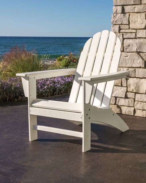 A white Adirondack chair positioned on a patio, with a scenic view of the ocean and lush plants in the background.