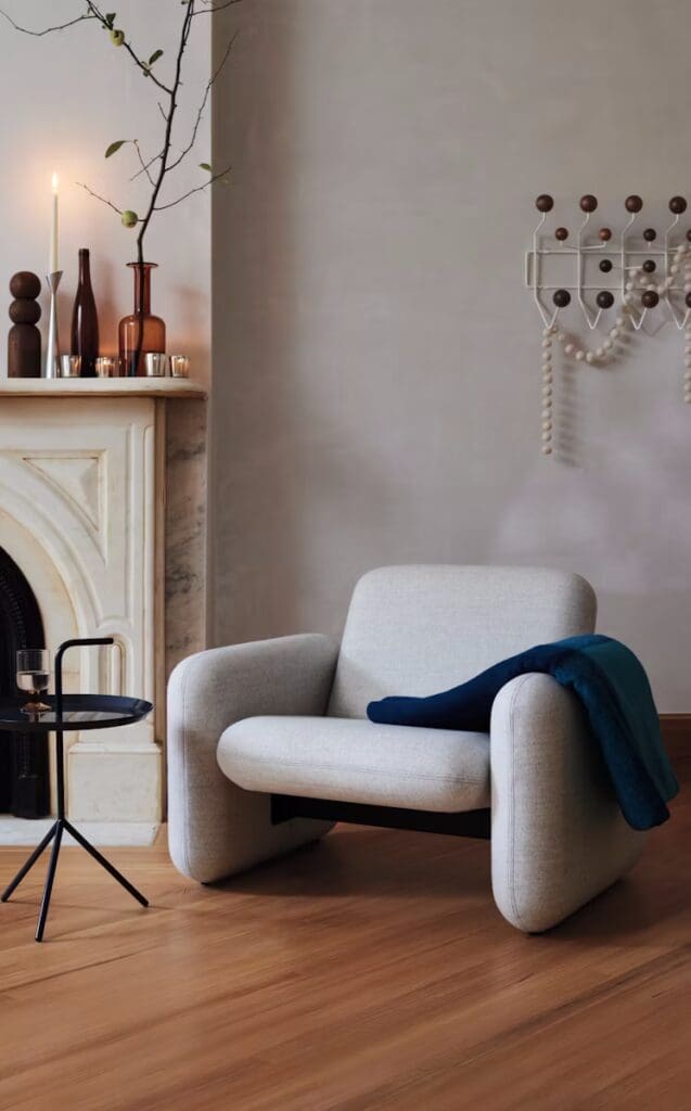 A modern armchair in a cozy living room setting, complemented by a black side table, a candle, and decorative vases on a marble mantelpiece.