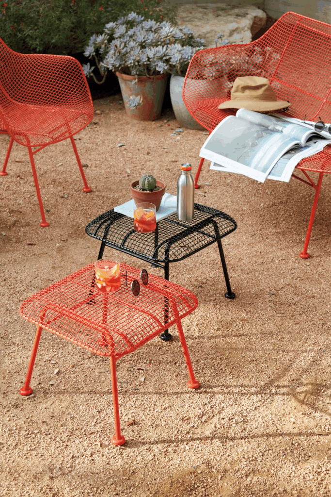 Colorful outdoor seating area featuring two red wire chairs and a black wire table on gravel, adorned with a cactus, a drink, a water bottle, and magazines.