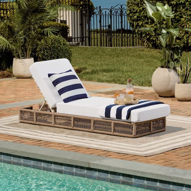 Stylish outdoor chaise lounge with a white cushion and navy striped accent, poolside on a brick patio, accompanied by a small side table with drinks and potted plants in the background.