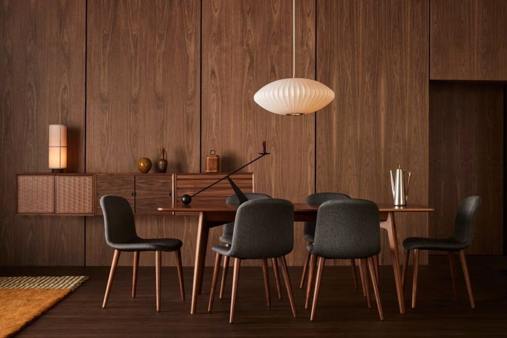 A modern dining room featuring a wooden table surrounded by gray upholstered chairs, with a cozy orange rug on the floor and warm wood paneling on the walls.